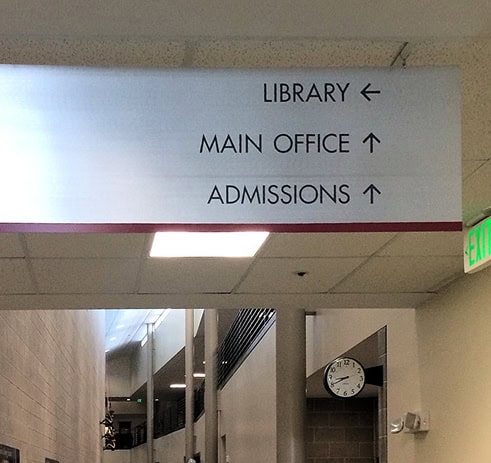 A hallway sign shows directions: Library to the left, Main Office and Admissions straight ahead. A clock and part of a hallway are visible below.