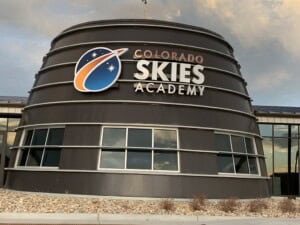 A modern round building with large windows and a sign that reads Colorado Skies Academy, featuring a space-themed logo with stars and an orange swoosh. Rocks and dried plants are in front of the building.