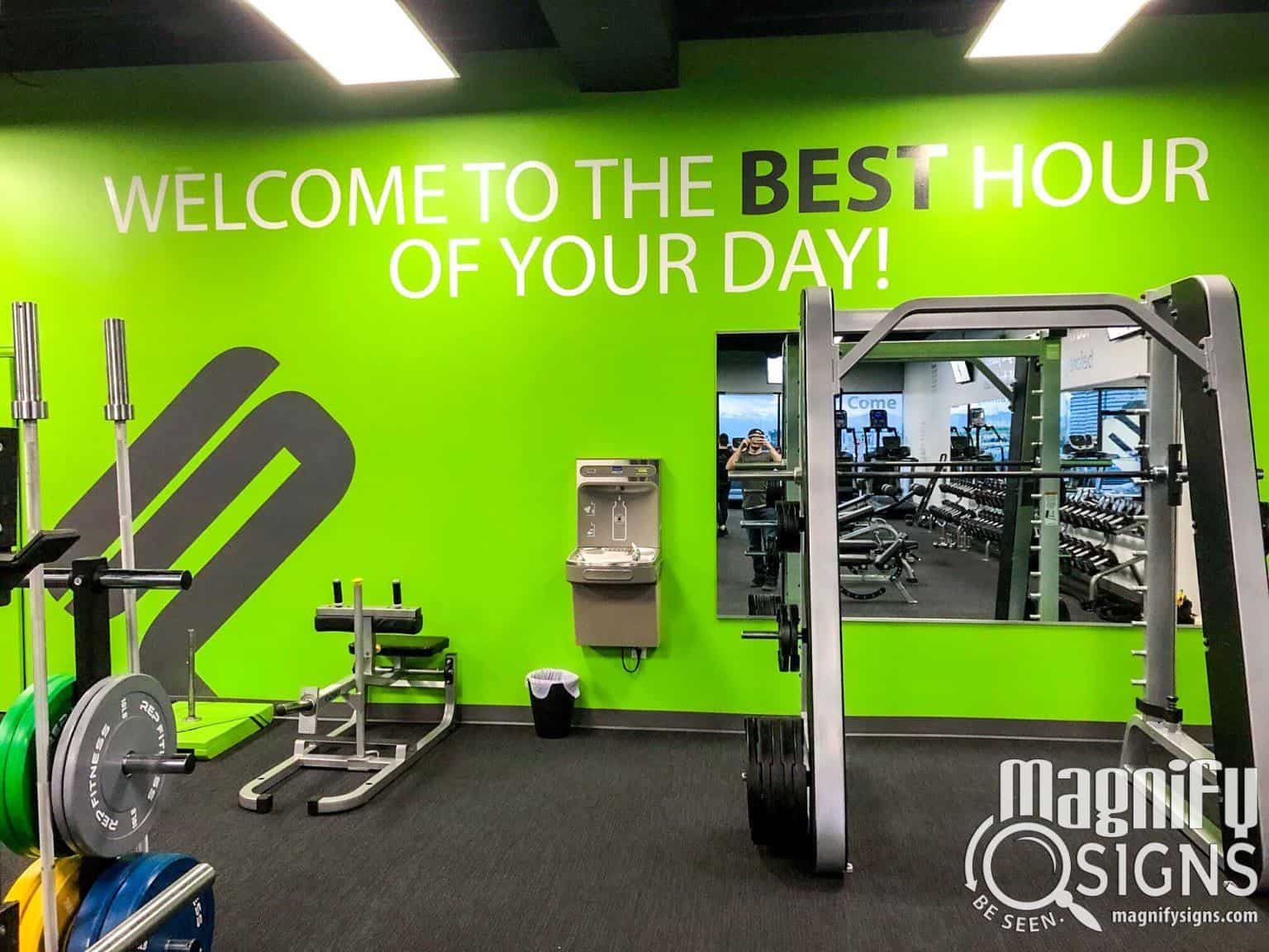 A gym with green walls, weightlifting equipment, a water fountain, and a wall sign that says WELCOME TO THE BEST HOUR OF YOUR DAY! The Magnify Signs logo is visible in the corner.