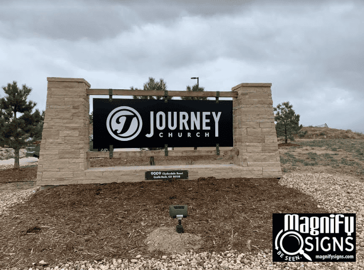A stone sign displays Journey Church with a circular logo, standing on a landscaped area with mulch and a small light. The Magnify Signs logo is in the bottom right corner. The sky is cloudy in the background.