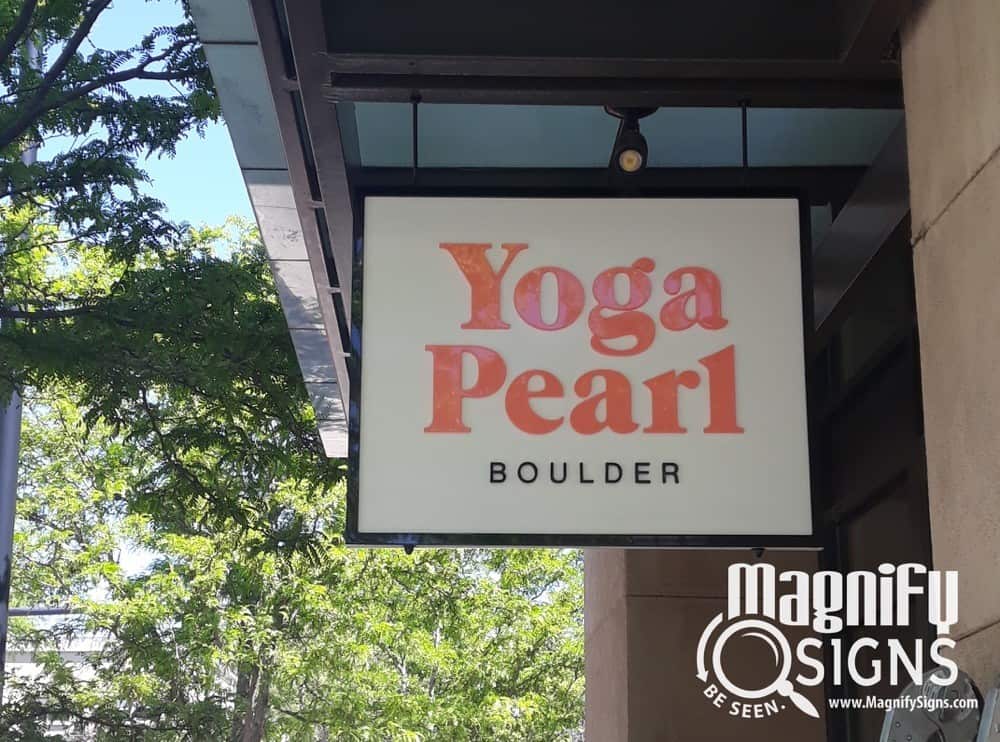 A hanging sign reads Yoga Pearl Boulder in large letters, with the Magnify Signs logo in the bottom right corner. Trees and a building are visible in the background.