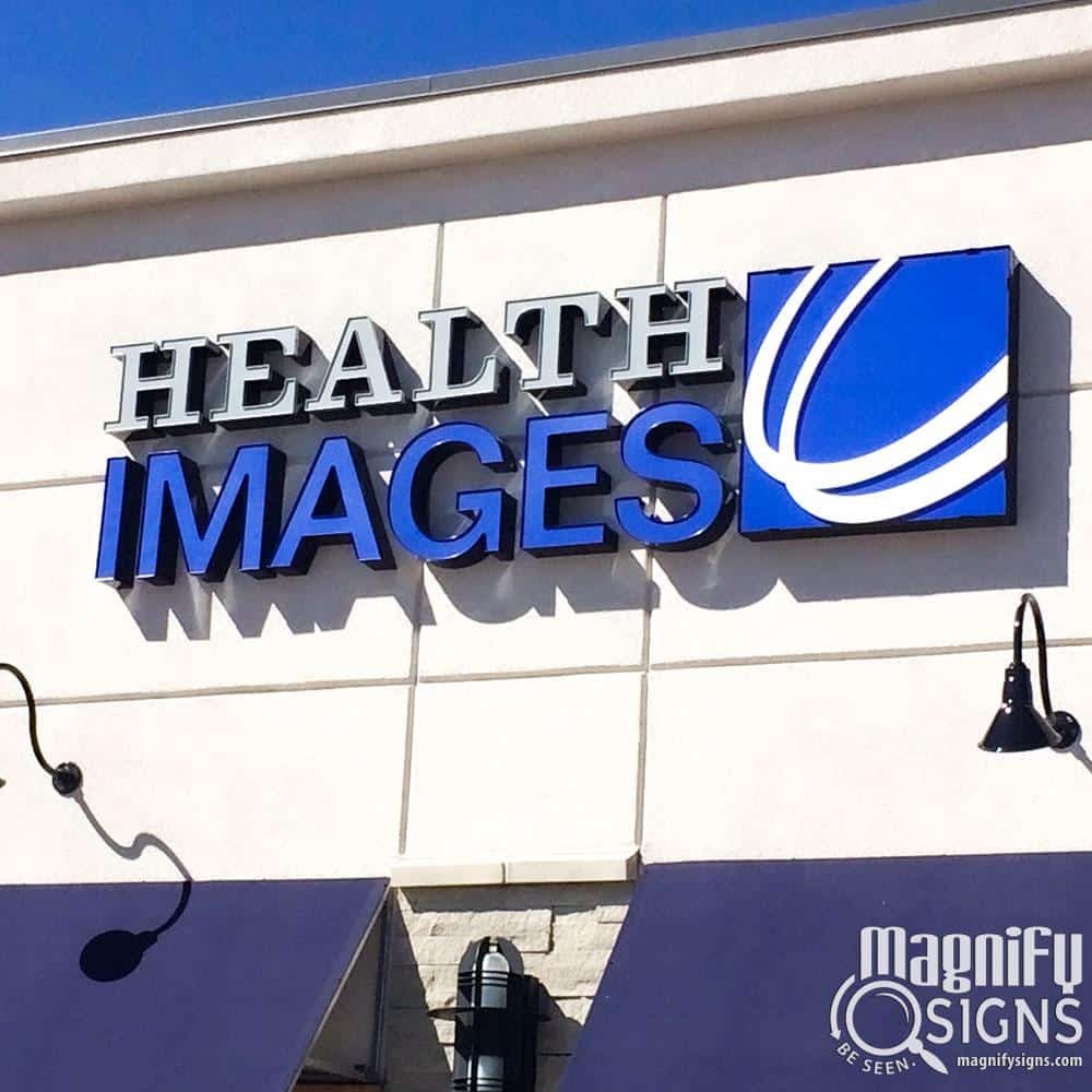 Storefront sign reading HEALTH IMAGES with a blue logo next to the word IMAGES on a light-colored wall. The sign is above a row of black gooseneck lamps. Magnify Signs logo is in the bottom right corner.