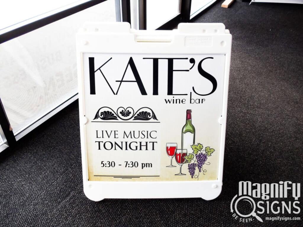 A sign for Kates Wine Bar advertising live music tonight from 5:30 to 7:30 pm, with wine and grape illustrations. The sign is indoors and branded with the Magnify Signs logo.