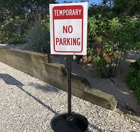 A sign on a black stand reads “TEMPORARY NO PARKING” in red and white. It is placed on a gravel path near a wooden barrier and green bushes.