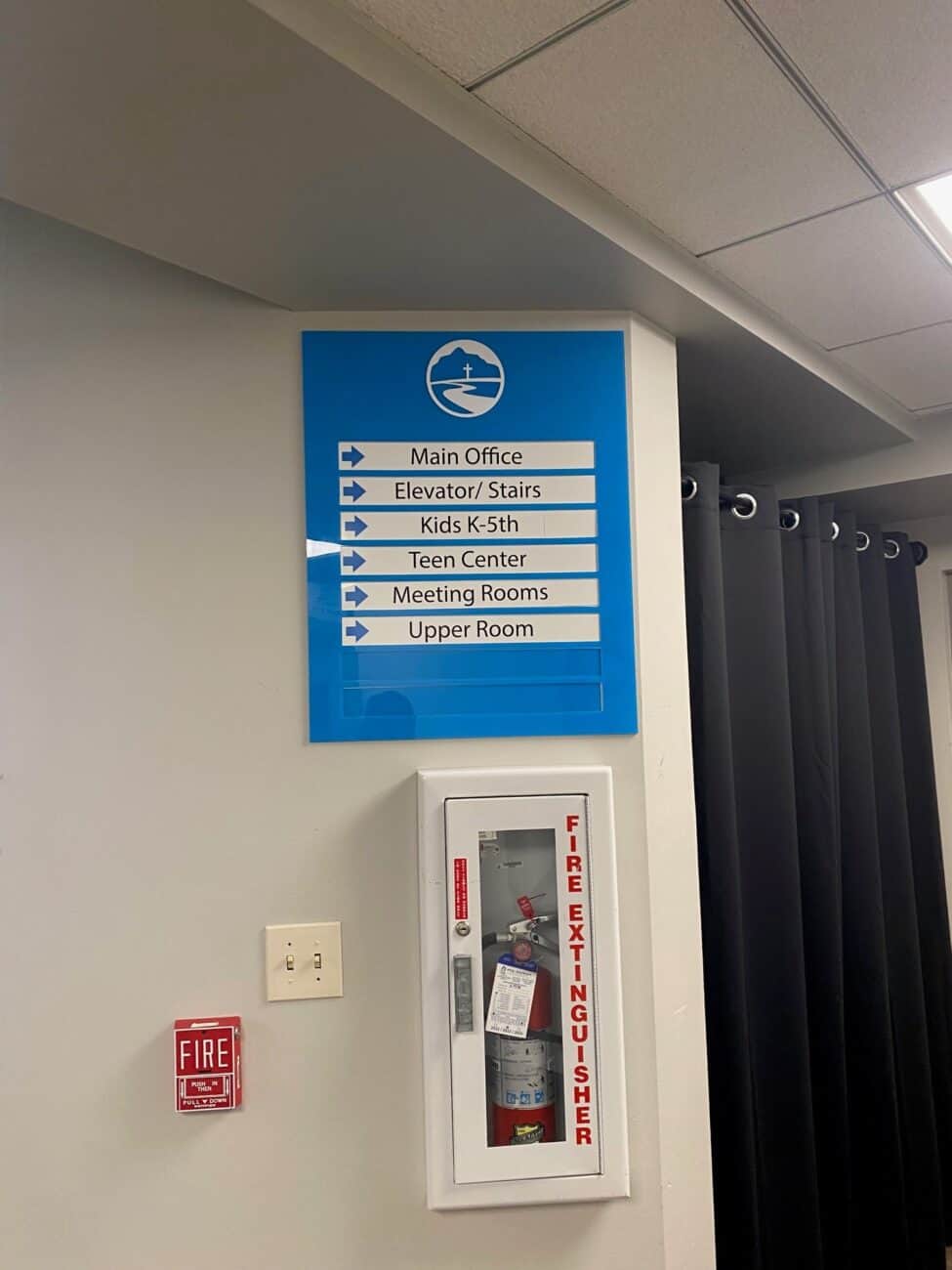 A blue sign with white arrows lists directions for Main Office, Elevator/Stairs, Kids K-5th, Teen Center, Meeting Rooms, and Upper Room. Below the sign is a wall-mounted fire extinguisher and fire alarm.