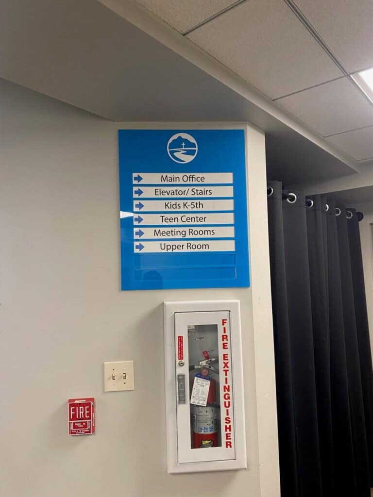A wall sign lists directions for Main Office, Elevator/Stairs, Kids K-5th, Teen Center, Meeting Rooms, and Upper Room. Below it is a fire extinguisher behind glass and a fire alarm on a white wall.