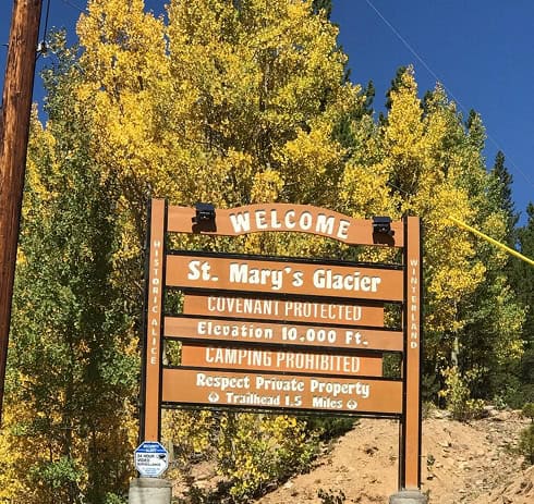 A wooden welcome sign for St. Mary’s Glacier stands in front of yellow autumn trees. The sign notes covenant protection, 10,000 ft elevation, camping prohibition, and mentions a trailhead 1.5 miles ahead.